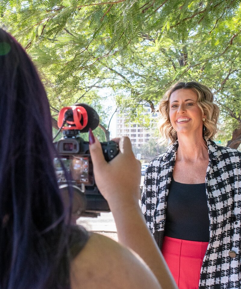 Smiling woman being filmed outdoors beneath green trees during a sunny Tucson interview session, photographed by Vyrl Photo, showcasing Tucson brand photography.