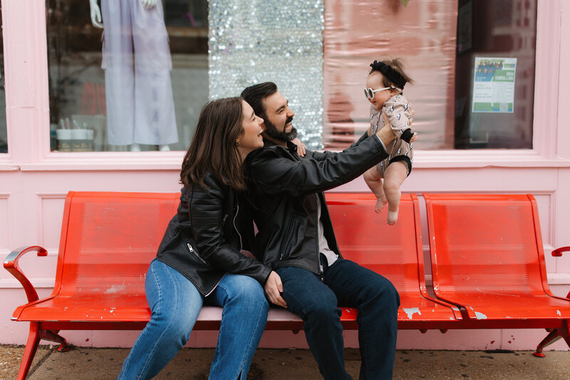 Mom and dad wearing leather jackets sit on red bench holding their infant daughter