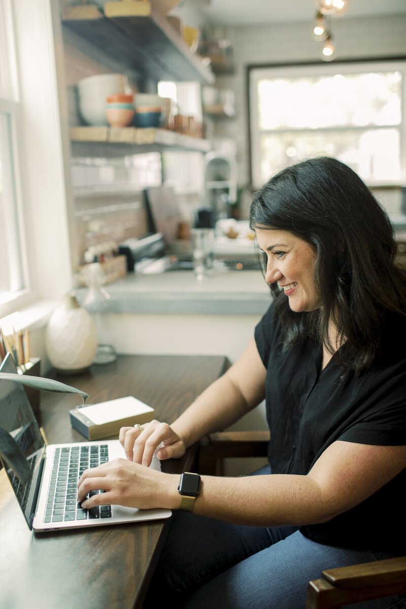 Online interior decorator, Michelle, sitting at a desk working on laptop wearing a black top and jeans