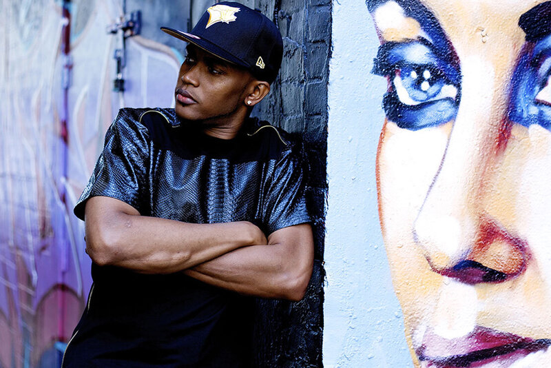 Musician portrait Fiyah MC leaning against black brick wall with graffiti of womans face beside him