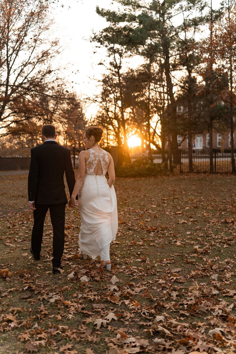 Bride and groom walking hand in hand toward the autumn sunset at Coxhall Mansion in Carmel, Indiana