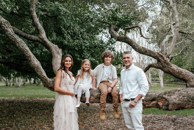 The Oosthuizen family sitting together on a large branch at the beach during their relaxed Auckland family photoshoot, captured by Zanthe Vorsatz Photography.