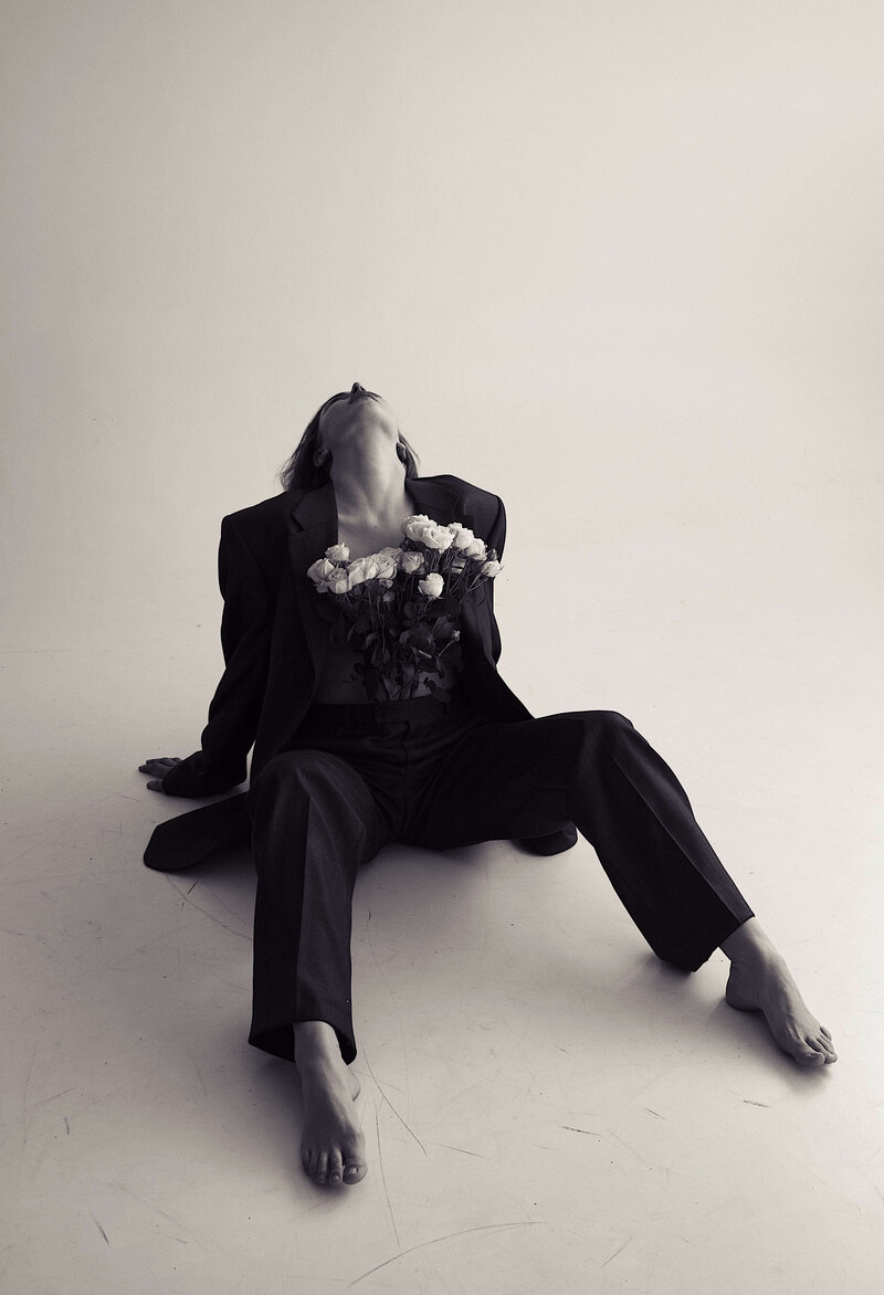 Black and white image of a brunette woman sitting on floor with head back in a black blazer with fresh flowers on chest