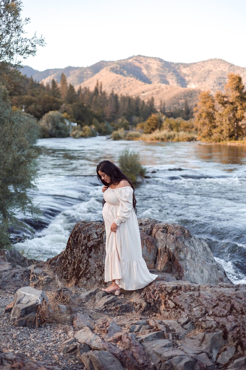 Lovely pregnant woman in cream flowy dress standing on rocks by the river