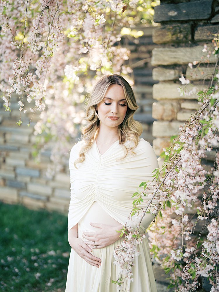 A blonde pregnant woman in a white dress stands next to a stone wall and cherry blossom tree in a DC garden during her maternity photos.