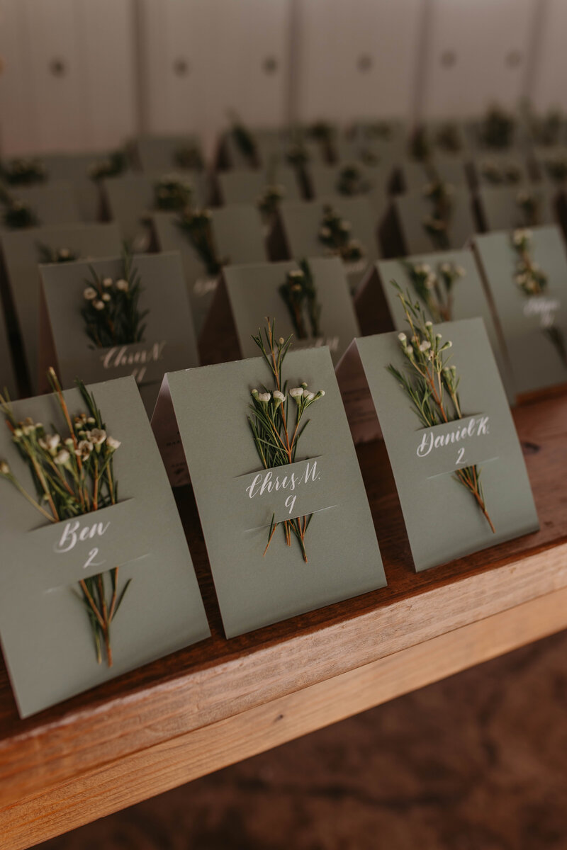 Wedding escort cards on sage green cardstock, each tied with sprigs of white florals.
