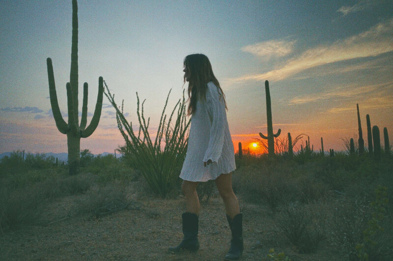A person in a white dress and black boots walks in a desert landscape with cacti at sunset, evoking the bold vision of a Sweet Pea Ink Creative web designer.