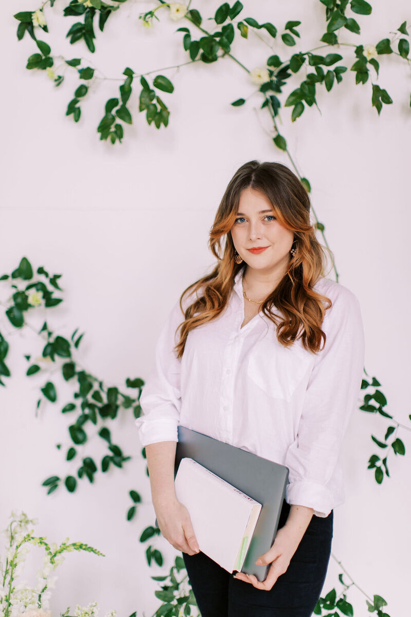 Amber looks at camera and stands in front of. a white wall with green vines  holding a laptop and book while wearing a white button down shirt and black pants