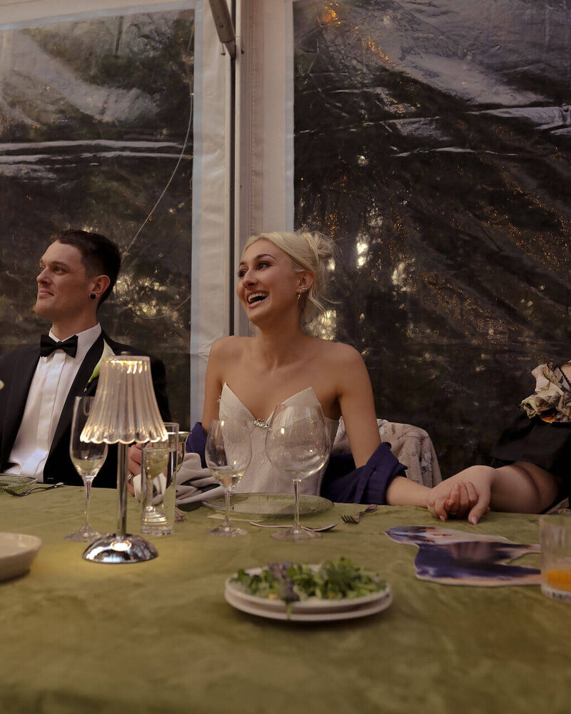 A bride laughs while seated at a table with a lit up LED lantern, representing luxury and affordable event rentals offered by Birdie June. 