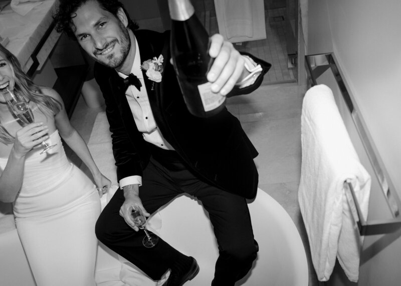 Black and white candid photo of groom holding bottle of champagne close to the lens with a glass of champagne in his other hand while bride takes a sip.
