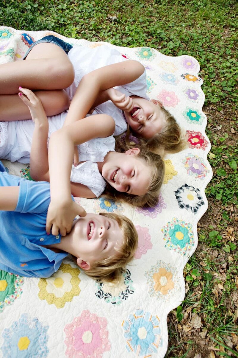 2 little girls and boy laying down on a colorful quilted blanket laughing together.