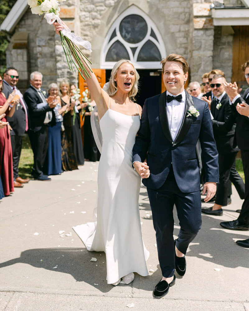 This cheerful photograph captures the bride and groom walking out of a church in downtown Calgary, with the bride’s arm in the air in celebration, surrounded by happy family and friends.