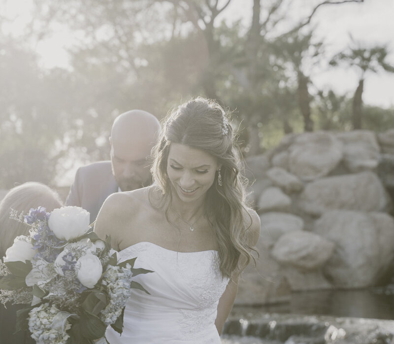 A bride, smiling gently, leads the way in a sunlit outdoor setting, holding a large bouquet of white flowers. Behind her, a groom follows closely. The scene is beautifully backlit, with soft sunlight filtering through trees, and a decorative rock waterfall adds a serene and romantic backdrop.