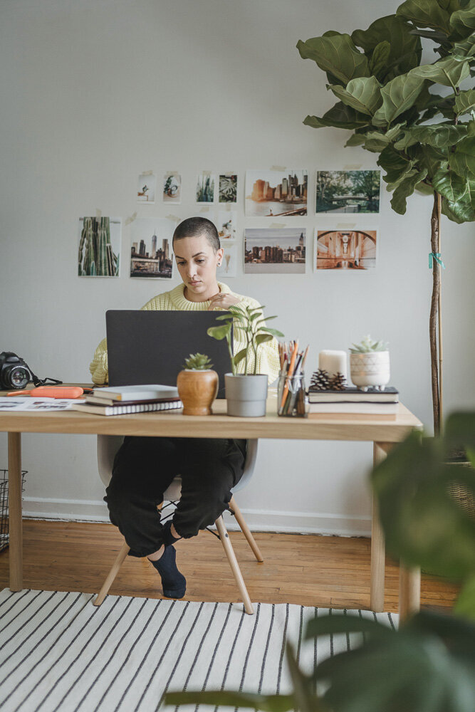 This image shows a feminine-presenting person with a buzzcut wearing a light green sweater and dark sweatpants. They are sitting at a desk full of books and houseplants, looking down at the laptop in front of them with a focused expression. The wall behind them is a collage of photos.
