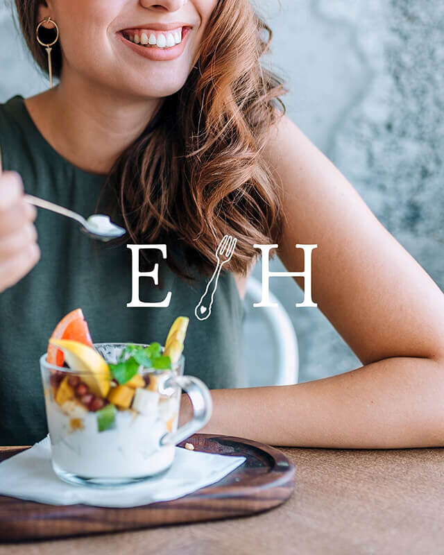 Stunning long-haired woman enjoying a delectable glass mug of creamy yogurt, topped with a colorful array of fresh fruits. With a radiant smile on her face and her hand poised delicately with a spoonful of yogurt, she gazes off into the distance. The image is overlayed with an example of a modern nutritionist logo.