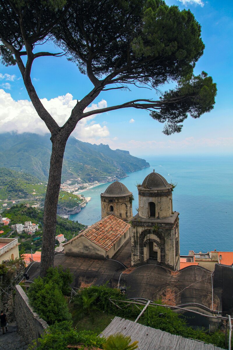 Panoramic view from Ravello overlooking the Amalfi Coast, with historic stone towers framed by a tall pine tree and the turquoise sea stretching into the horizon.