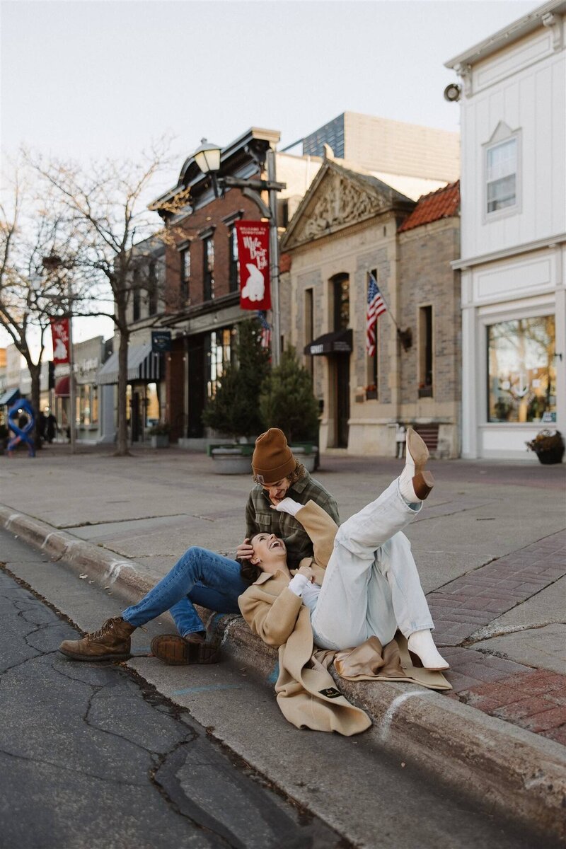 Woman lays on street curb with her head in her fiance's lap during small town Minnesota engagement session