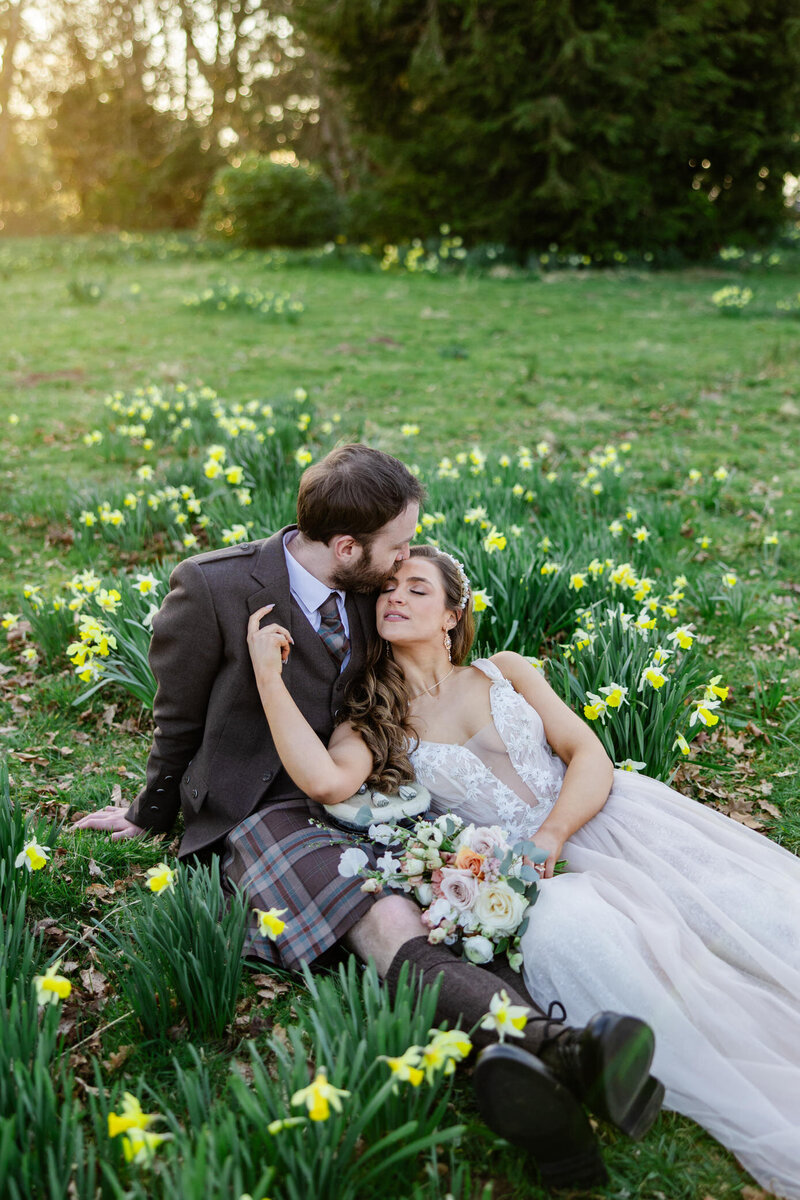 kilt wearing groom kisses bride in field of daffodils