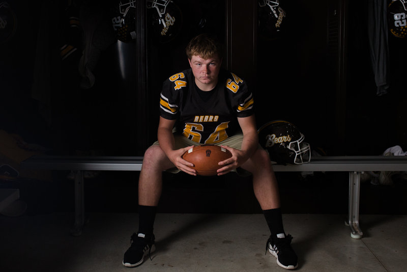 High school senior guy in locker room, sitting on bench holding football wearing jersey and shorts, dimly lit room with dramatic lighting, photographed by Jamie Lynette Photography