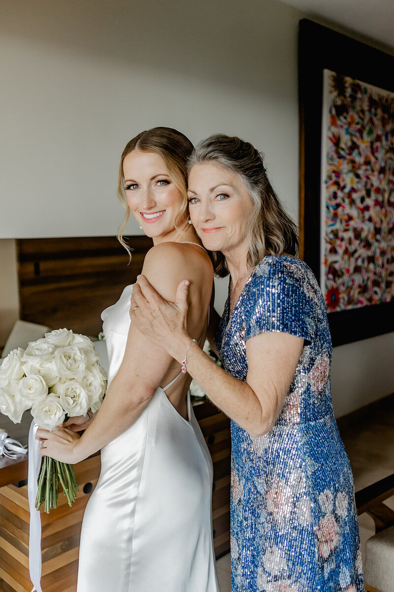 A bride stands next to her mother in a heartfelt pose before the wedding ceremony. The bride holds a bouquet of white roses, and the mother wears a sparkling blue dress as they both smile at the camera