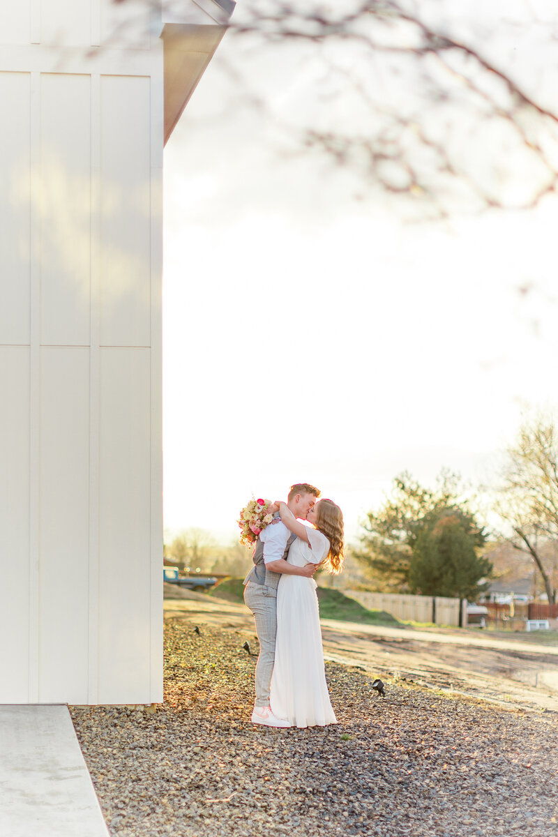 Couple kissing on the side of white barn.