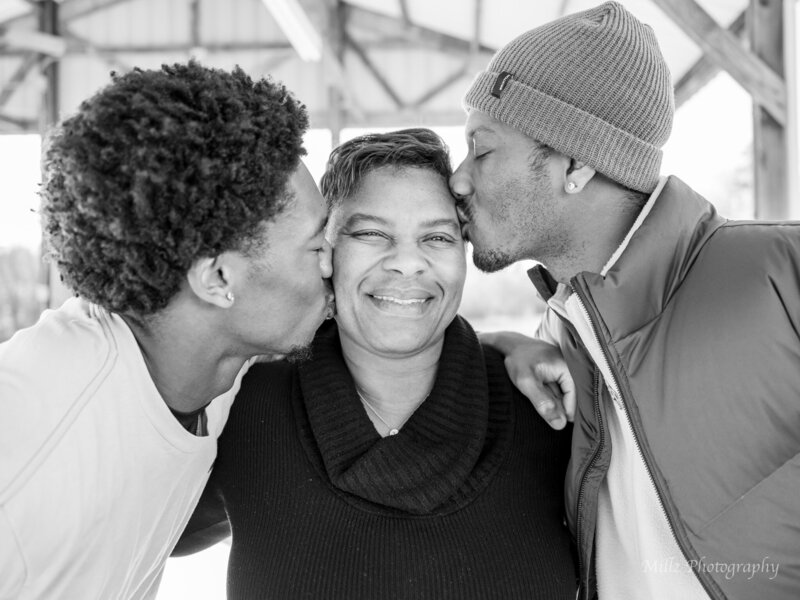 a black and white photo of a mother with her two grown sons, each giving her a kiss on the cheek. photographed by Millz Photography in Greenville, SC