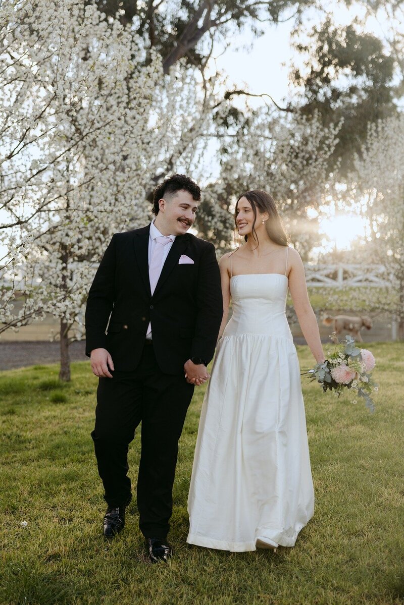 A bride in a strapless dress walking with her groom thrugh white cherry blossoms.