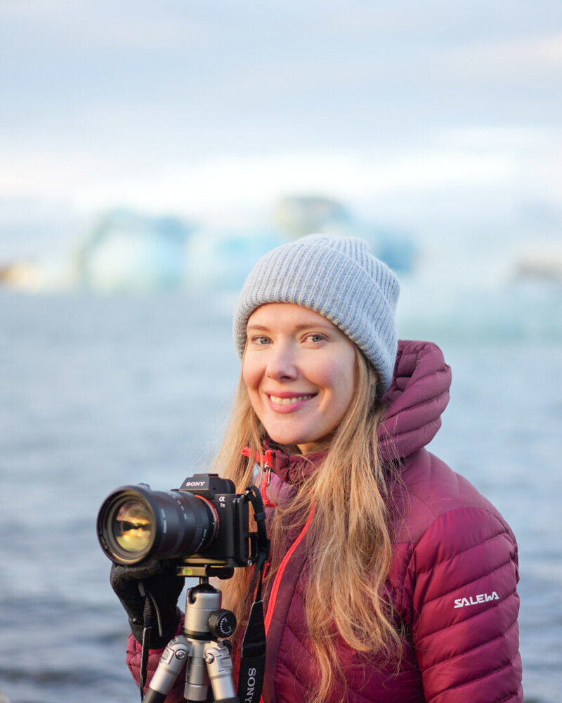 Anna Isabella Christensne - a female photographer smiling and looking into the camera next to her camera set up on a tripod. She is sitting next to Jokulsarlon glacier lagoon in Iceland.