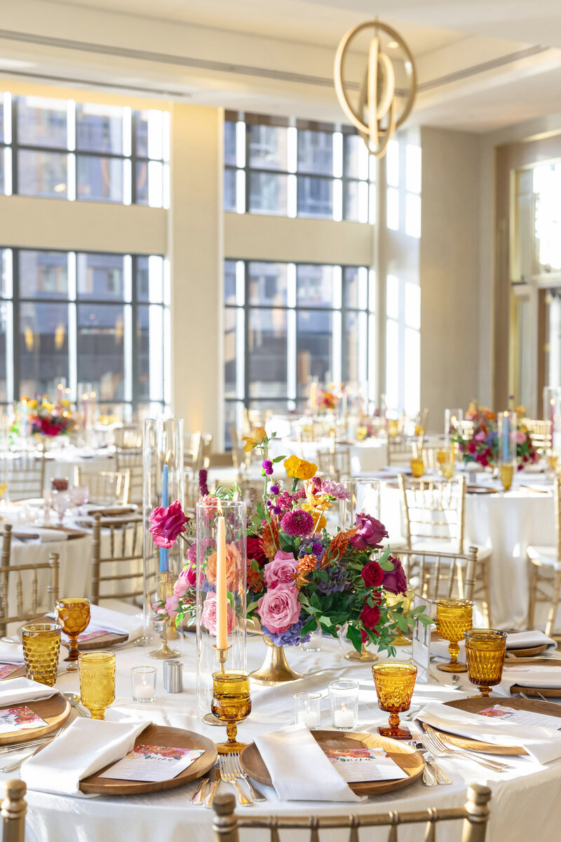 A brightly lit ballroom featuring elegantly set tables with gold chairs. Each table has colorful floral centerpieces, gold-rimmed plates, and amber glasses. Oversized windows bring in natural light, creating a warm atmosphere.