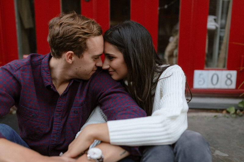  A couple sits on the ground in Hoboken, framed by a bright red door, capturing a cozy and intimate moment.