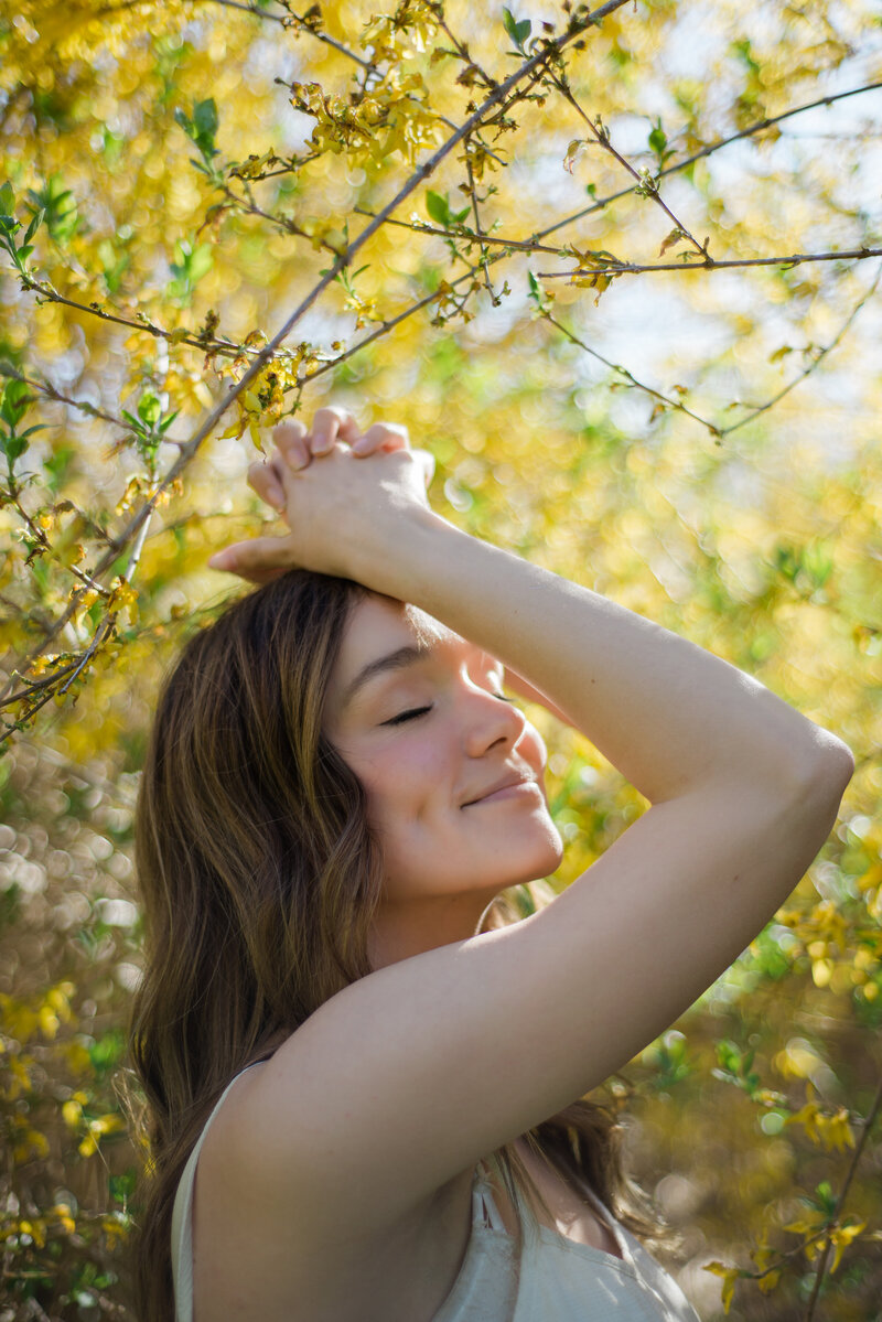 Woman smiling and closing her eyes with her arms over her head in front of yellow flowers
