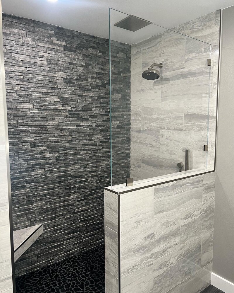 A remodeled shower with a dark grey thin tiled wall and a pony wall with a half glass enclosure on the other side.