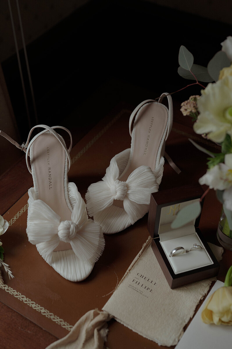 Bröllopsskor, vigselringar och inbjudan står tillsammans på ett bord.