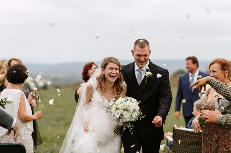 A newlywed couple walks joyfully down an aisle, petals falling around them, surrounded by smiling guests on a cloudy day.