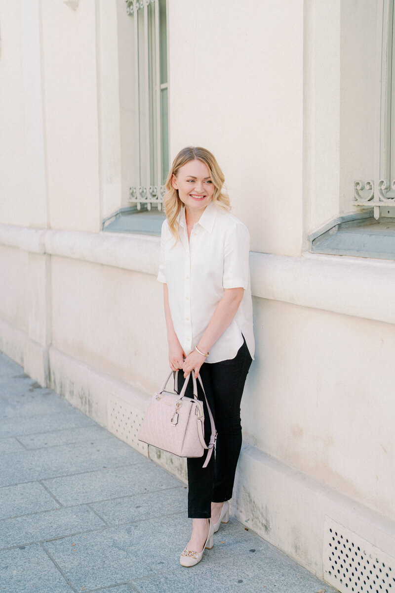 a woman in a white top and black pants  leaning up against a white building in Paris holding a hand bag and smiling while looking off into the distance