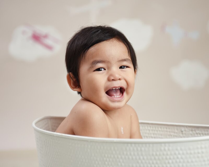 A baby boy laughing while sitting in a white tub, with a cloud and airplane-themed background.