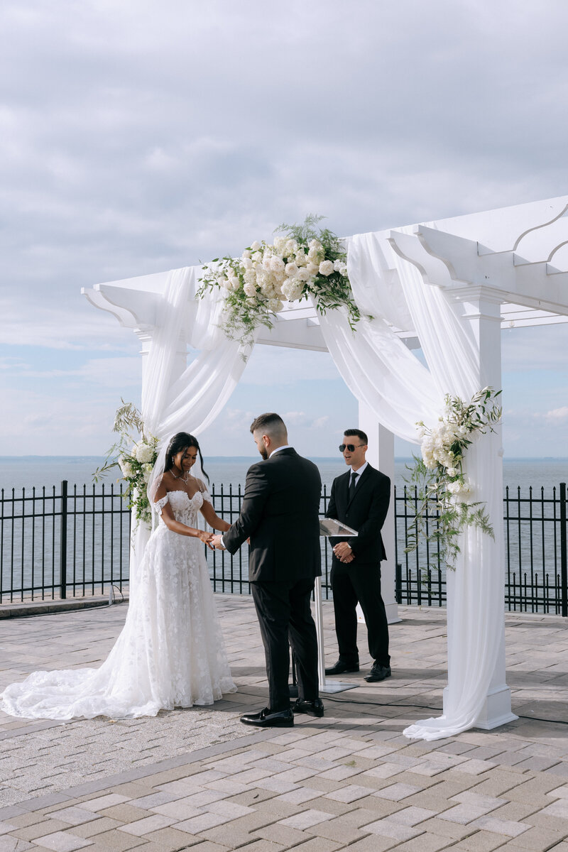 Bride and groom exchanging vows during outdoor wedding ceremony under a white floral arch by the water in New York, with elegant draping and ocean view backdrop