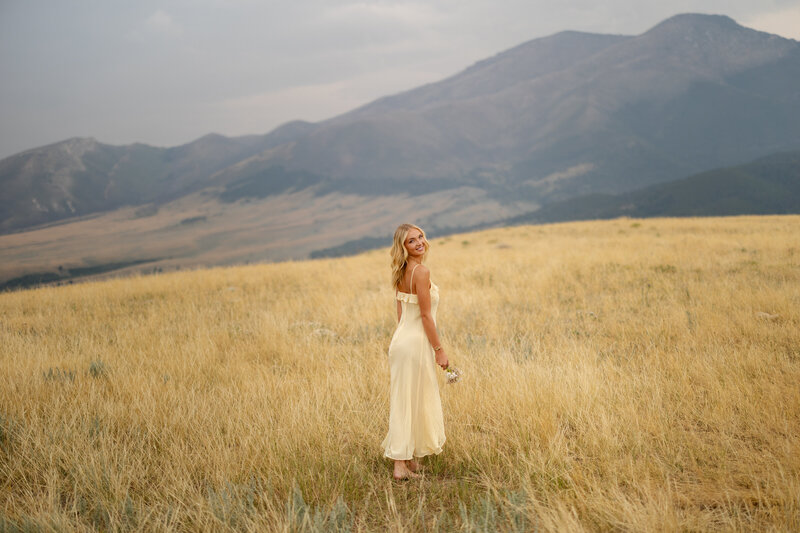 High school senior smiles as she stands in the middle of a wildflower field tree-lined mountainsides.
