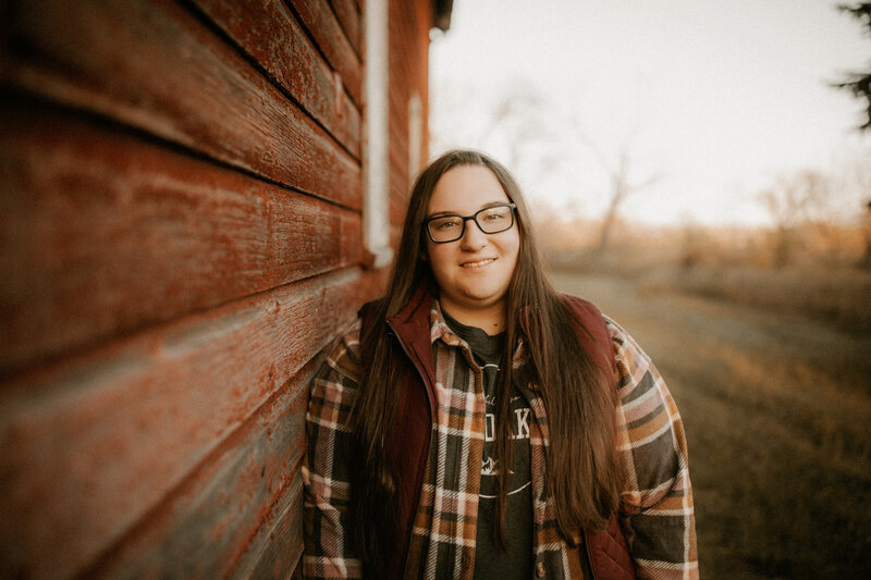 Photo of a senior girl leaning up against an old barn and smiling at the camera.