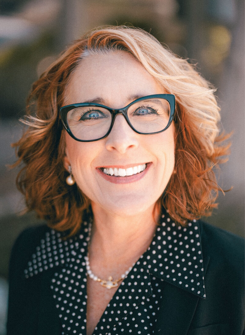 Portrait of Carolynn Santaniello smiling outdoors, wearing glasses and a black polka dot blouse with a blazer.