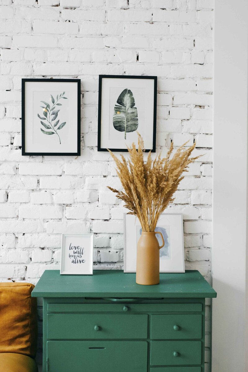 vase of dried grasses on top of a green dresser in front of a whitewashed brick wall