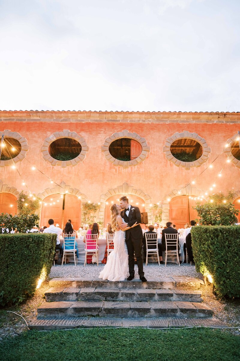 Bride and groom kissing in front of a warmly lit Italian villa courtyard surrounded by guests at a long dinner table under string lights.