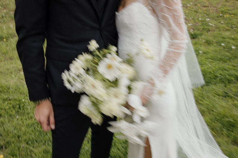 couple in black suit and white wedding dress lean close together while holding a white and green floral bouquet