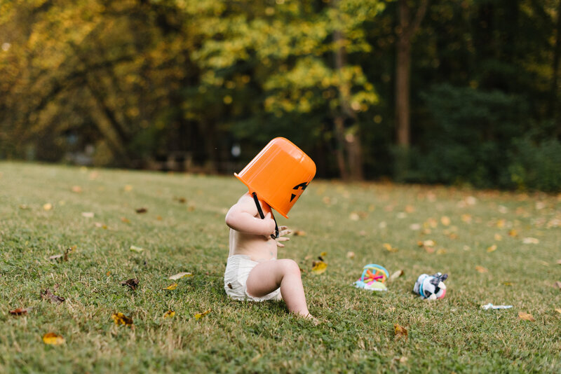 Baby in diaper wearing a pumpkin bucket on head sitting on grass - Washington DC Family Photographer