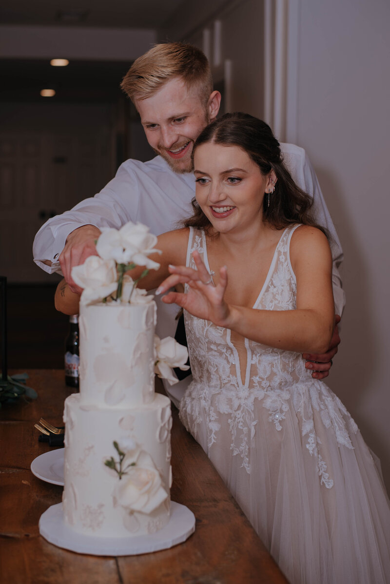 The bride and groom beam as they cut a two-tiered white wedding cake adorned with delicate white flowers. The bride's lace dress and the groom's crisp white shirt perfectly capture the charm of a rustic wedding venue, beautifully depicted in photos from our Leesburg, VA venue gallery.