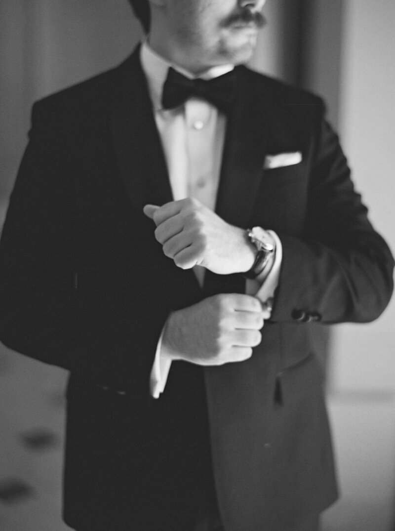 Newport Wedding Photographer | A man in a classic black tuxedo and bow tie adjusts his cufflinks in a dimly lit room.