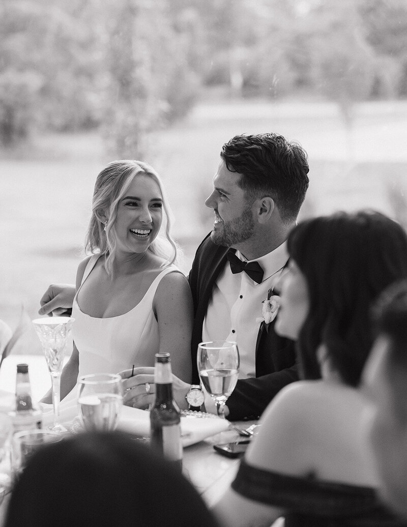 A smiling Ohio couple shares a joyful moment at an elegantly set table with drinks, surrounded by wedding guests.