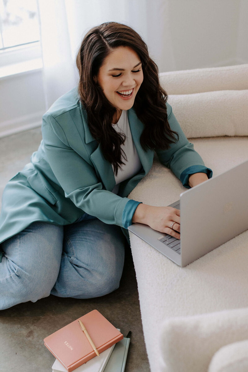 woman with dark hair wearing teal blazer on her laptop on floor
