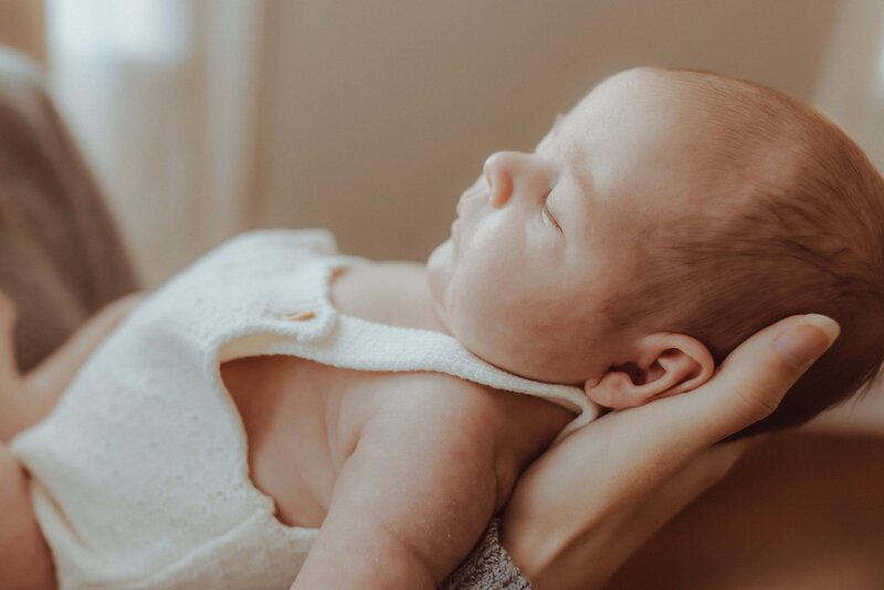 Close-up of a sleeping baby in a white outfit, cradled in someone's arm, against a softly lit background.