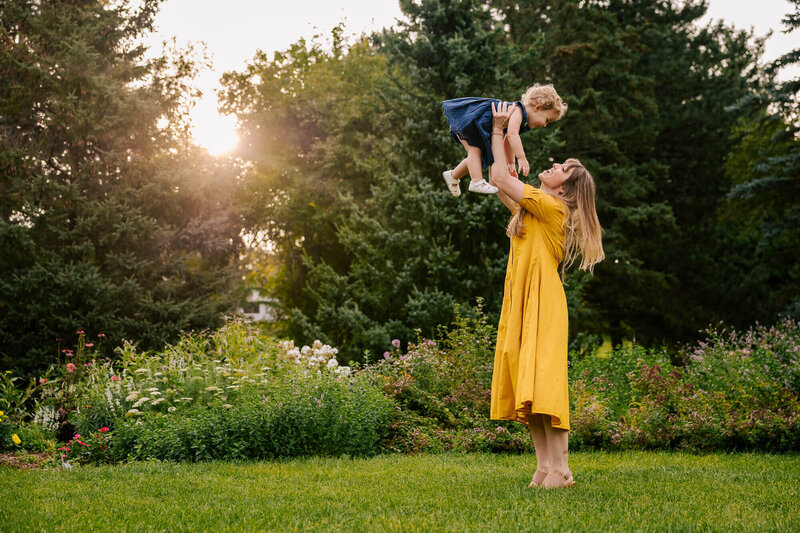 A young mother in a yellow sundress holds her daughter high into the air. The daughter has golden ringlets and she's smiling down at her mother. The sun peaks through the trees behind them and they're surrounded by flowers.